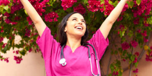 Smiling nurse in pink scrubs raises her hands to the sky while standing in front of a flowering shrub.