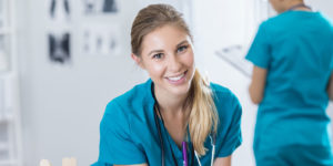 Young nurse smiles at the camera while her colleague reviews a patient chart in the background.